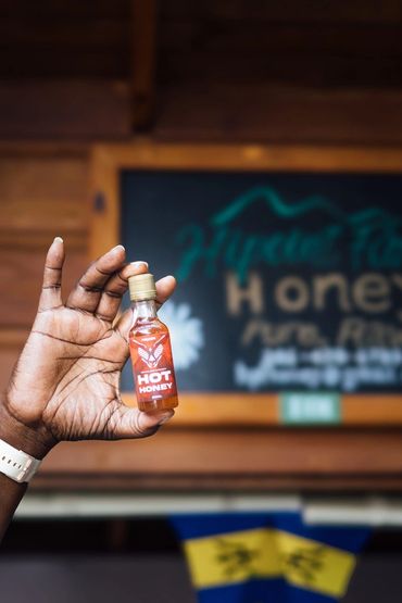 Hand holding a small bottle of Hipoint Farm hot honey sauce with a wooden background at Haymans Market Barbados store..