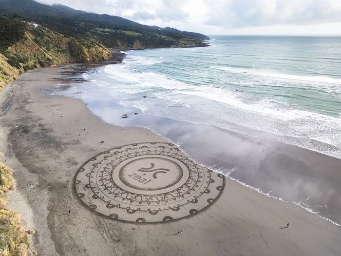 Intricate circular sand art with the word 'zeal' on a beach by the ocean.