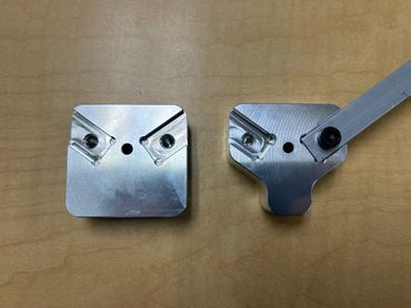 Two machined metal parts on a wooden surface, one with an attached metal arm.