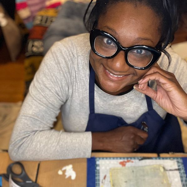Smiling woman wearing large glasses and an apron at a crafting table.