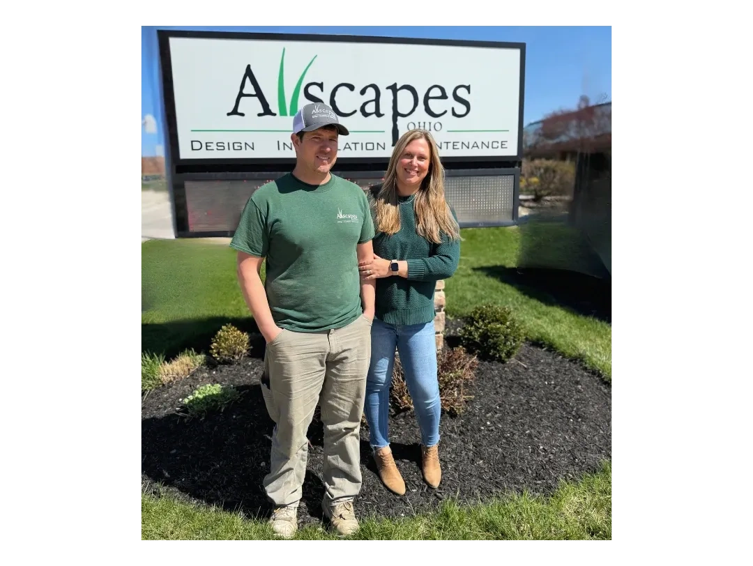 Two people standing in front of a landscaping business sign on a sunny day.