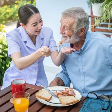 A caregiver joyfully feeding an elderly man outdoors at breakfast.