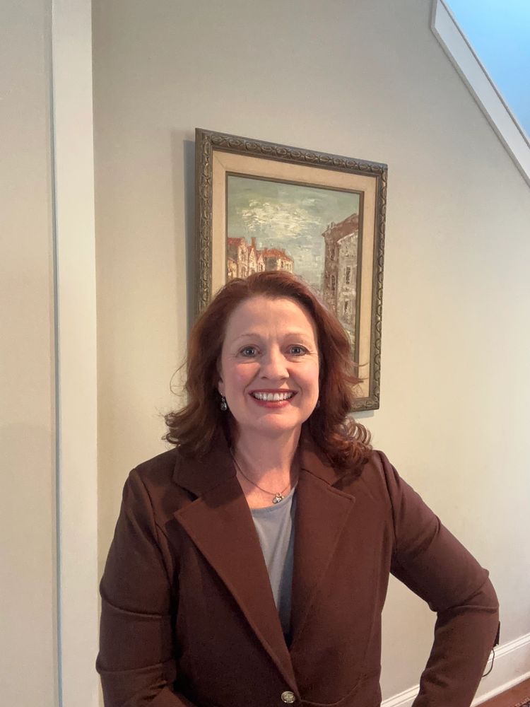 Smiling woman in a brown blazer stands indoors with a framed painting behind her.