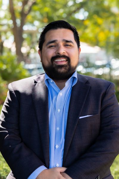 Smiling man in a blue shirt and dark blazer outdoors.
