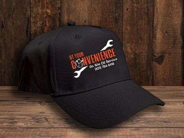 Black hat on a wood background. Hat has a logo for At Your Convenience Oil Service on it/
