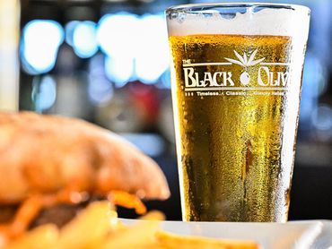 Beer glass with The Black Olive's logo on it behind a burger in the foreground