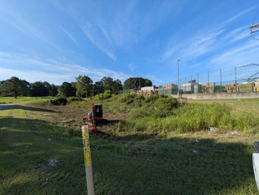 Small excavator working on grassy land near fenced storage area under blue sky.