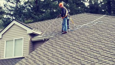 A man wearing safety gear cleans a residential roof with a power washer.
