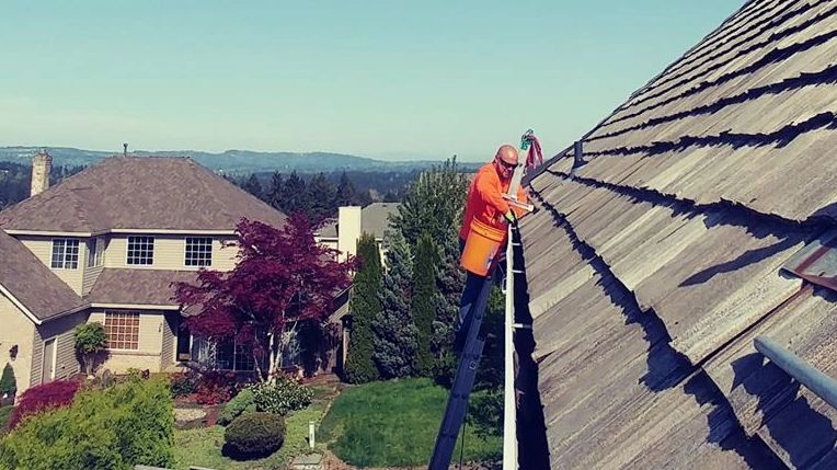 Worker on a ladder cleaning the gutter along a steep roof.