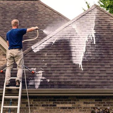 Worker soft washing a roof, gently removing dirt and grime with specialized equipment.