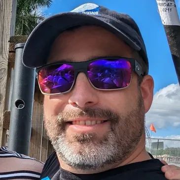 Man wearing reflective sunglasses, a cap, and a slight smile outdoors.