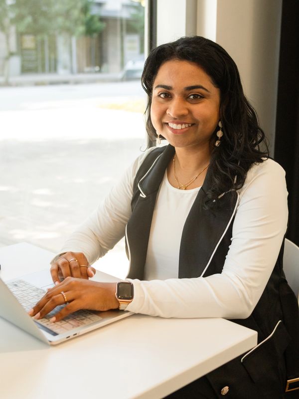 A smiling woman working on a laptop at a bright desk near a window.