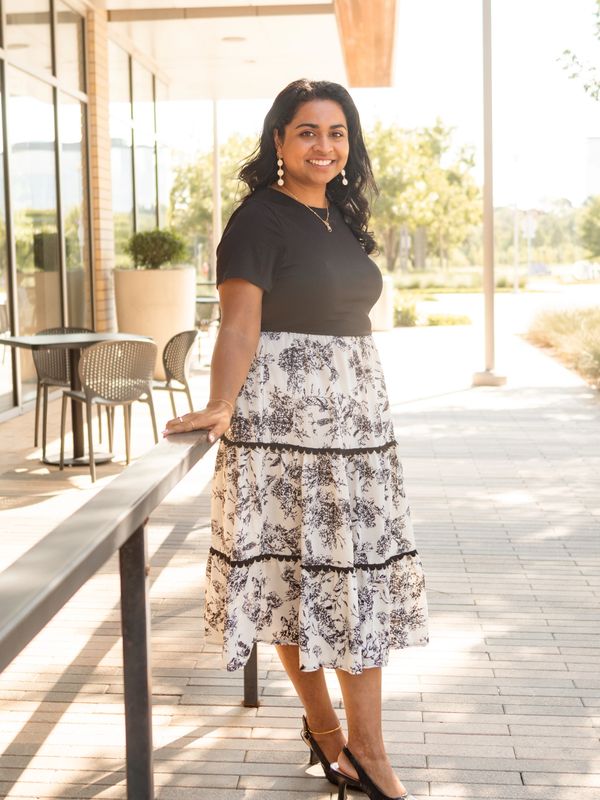 Woman in a black top and floral skirt smiling outdoors on a sunny day.