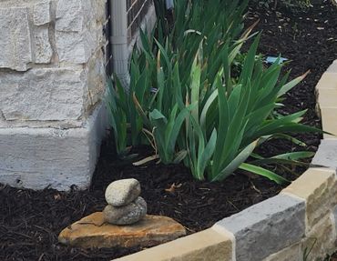 Stacked stones on a rock beside green plants and a stone wall.