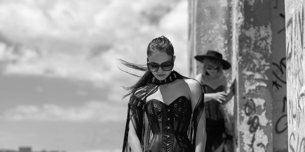 Black-and-white editorial portrait of two models in corsetry beside a weathered concrete structure.
