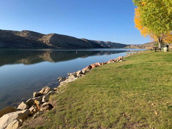 Calm lake with rocky shore and autumn trees under clear blue sky.