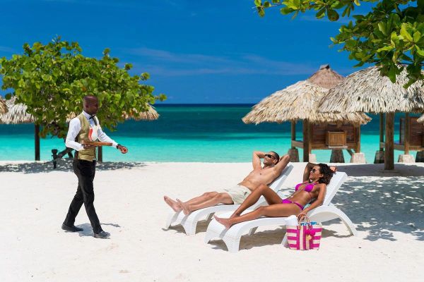 Couple relaxing on beach loungers with a waiter approaching.