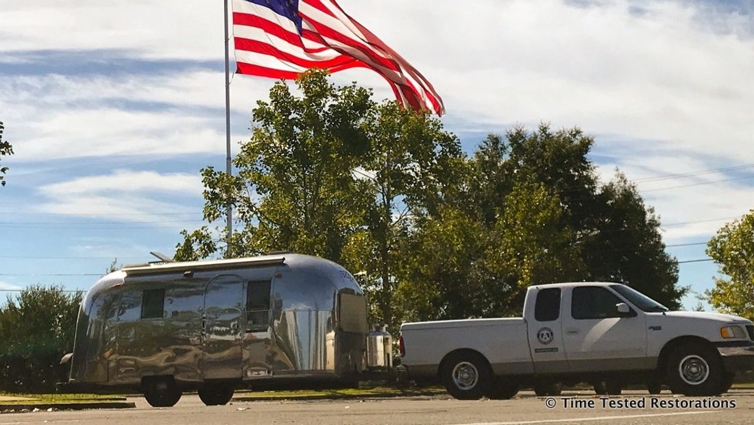 A shiny Airstream travel trailer and pickup truck under a very large American flag.