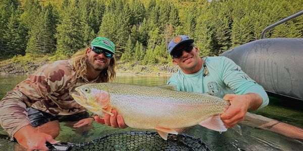 Two men proudly display a large rainbow trout by a river.