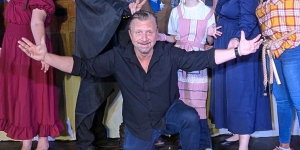 A diverse theater cast poses joyfully in costume on stage with a painted Western backdrop.