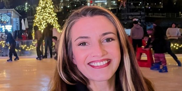 A smiling woman ice skating at night with festive Christmas lights in the background.