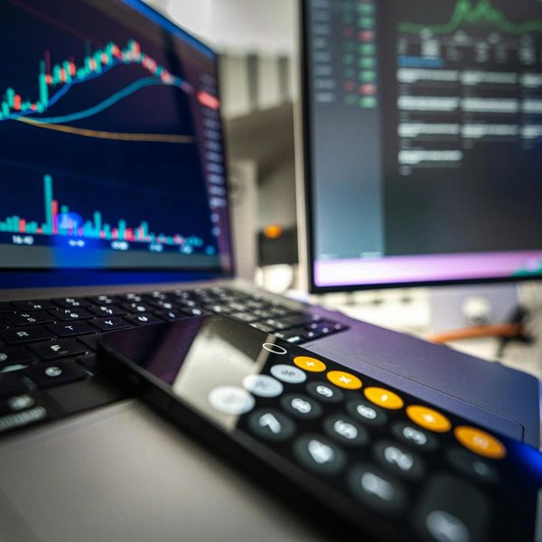 Calculator on a laptop with stock market charts on screens.