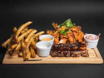 A wooden platter with ribs, fries, fried chicken, sauces, and coleslaw.