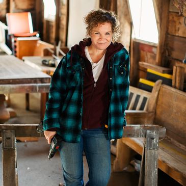 Woman in a workshop, smiling, holding a drill, leaning on sawhorse.