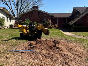A stump grinder machine and wood chips in a yard with houses in the background.