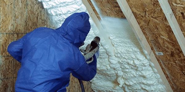Person sprays foam insulation between wooden beams in an attic.