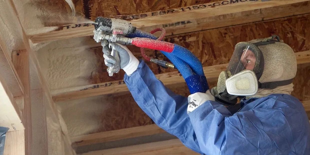 Worker sprays insulating foam on wooden ceiling beams, wearing protective gear.