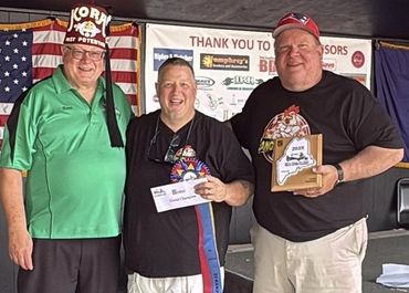 Three men smiling, holding awards at an event with an American flag backdrop.