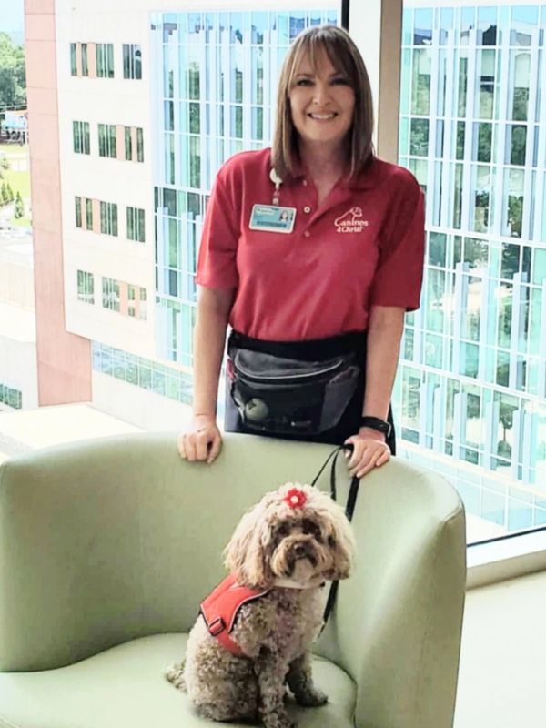 A woman in red with a therapy dog wearing a red vest and flower accessory.