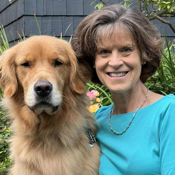 Woman in blue shirt smiles beside a golden retriever in a garden.