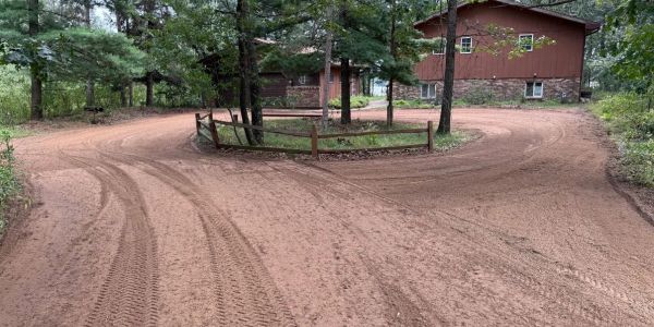 Dirt road splits around a fenced tree island near a house.