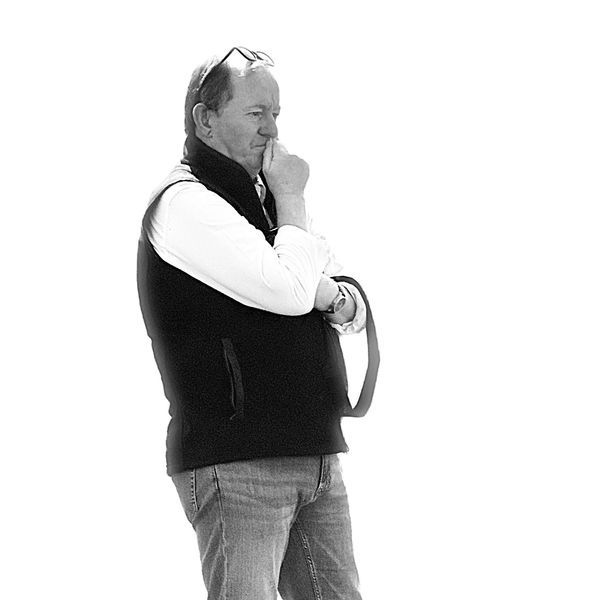 Man in black vest looks thoughtful with hand near mouth.