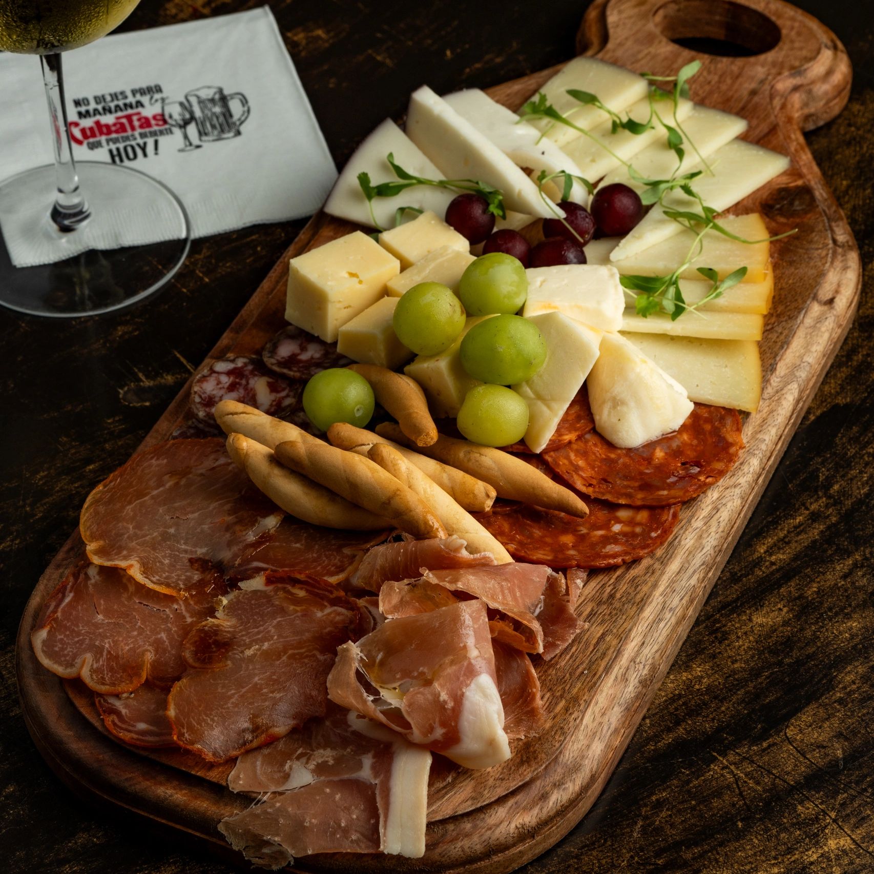 Charcuterie board with assorted cheeses, cured meats, grapes, and breadsticks alongside a glass of white wine.