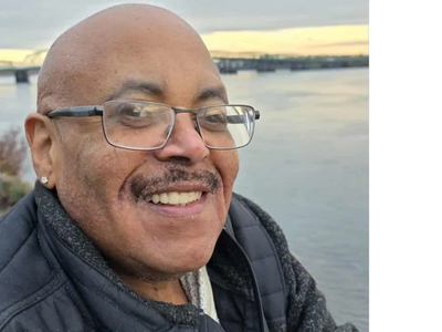 Jerry Woodards, smiling wearing glasses near a water body during sunset.