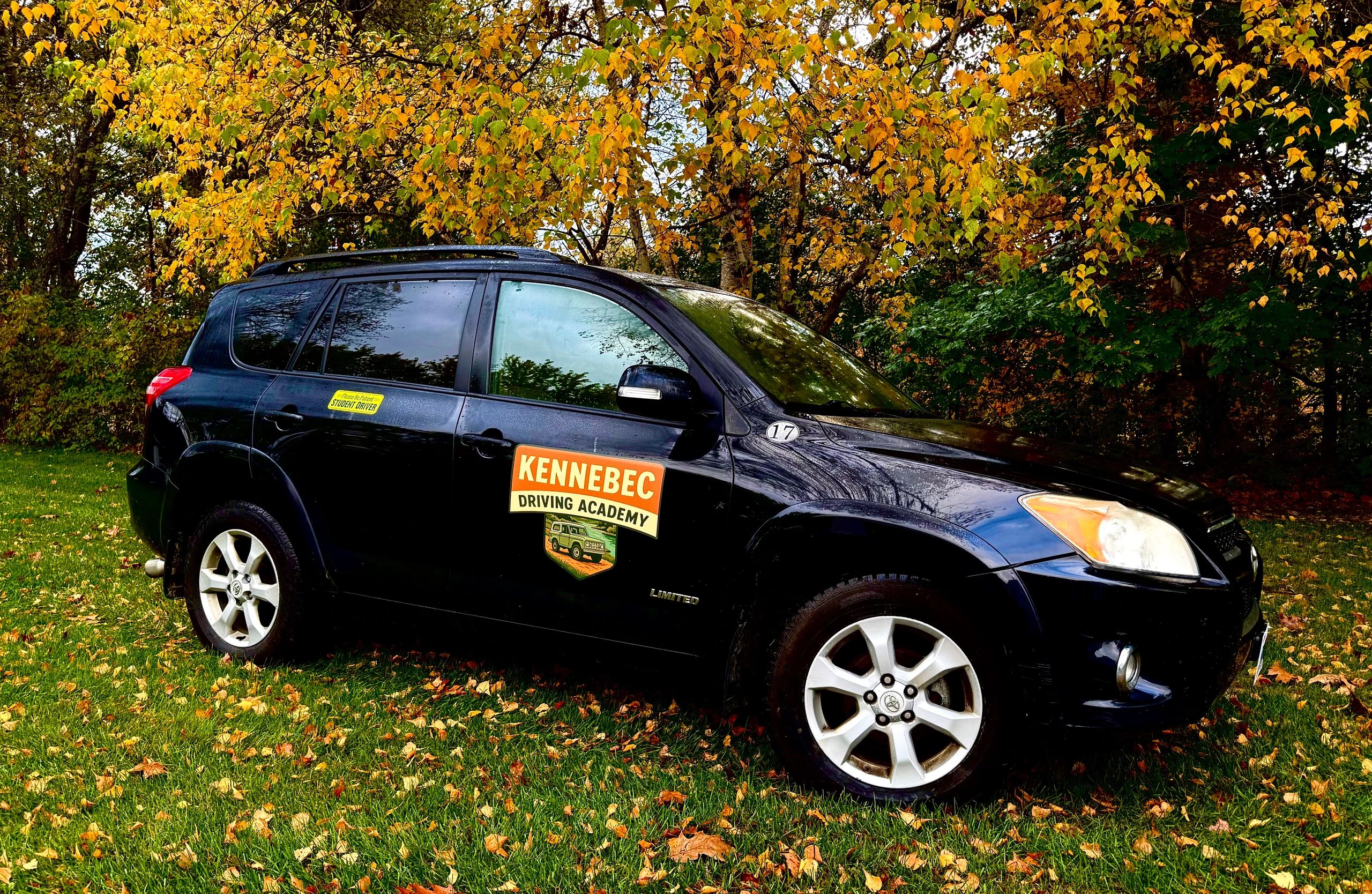 Black SUV from Kennebec Driving Academy parked on grass with autumn trees.