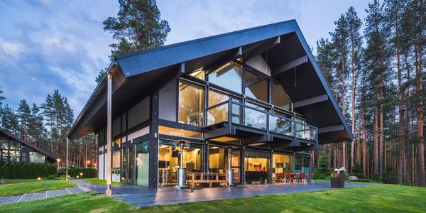 Modern glass house with a sloped roof surrounded by pine trees at dusk.