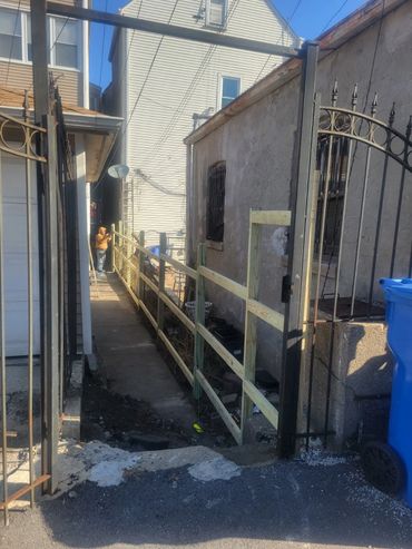 Two people working on a narrow alley fence between houses on a sunny day.