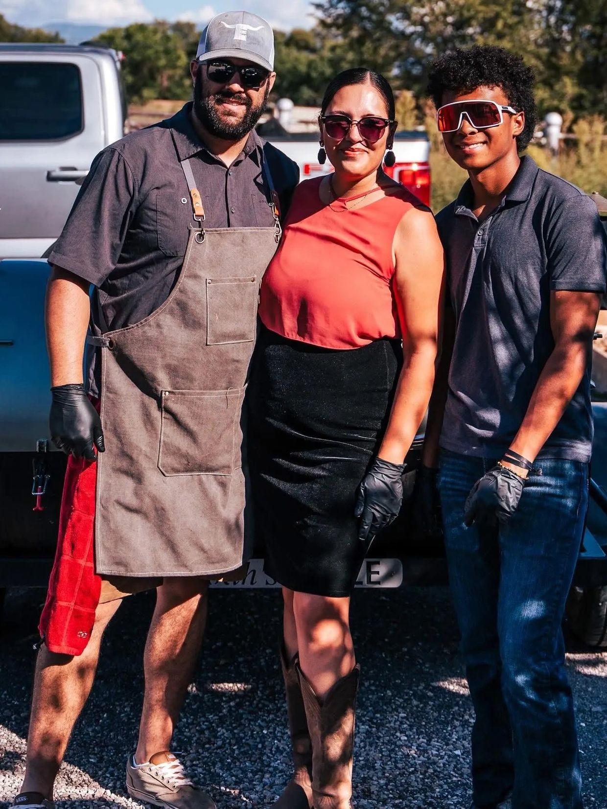 Three people smiling outdoors, wearing sunglasses and gloves.