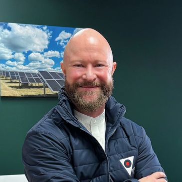 Smiling bald man with beard in a navy jacket, standing confidently indoors.