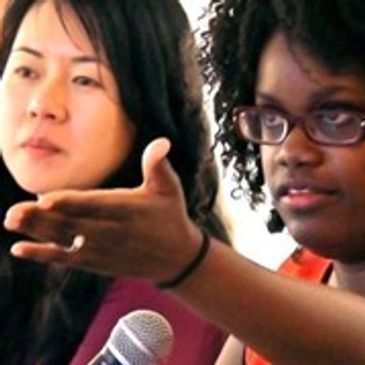 Two women in discussion, one gesturing while speaking into a microphone.
