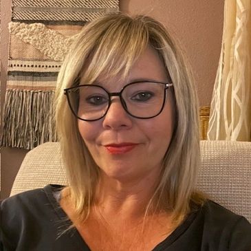 Smiling woman with blonde hair and glasses sitting indoors.