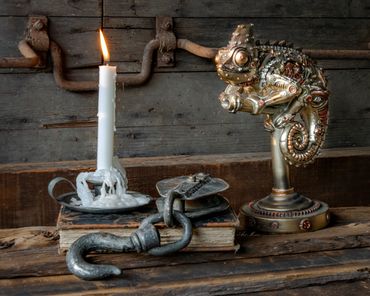 A lit candle, an old book with a metal hook, and a steampunk chameleon sculpture.
