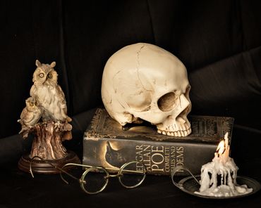 Still life with skull, Edgar Allan Poe book, owl figurine, glasses, and burning candle.