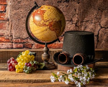 Vintage globe with steampunk hat, grapes, and white flowers on rustic wooden table.