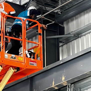 A worker in a blue vest and hard hat on an orange lift working on ceiling pipes.