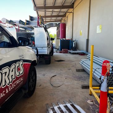 Work trucks and metal pipes stored in an industrial area under a corrugated roof.